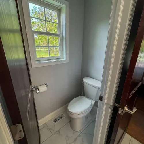 Brightly lit bathroom featuring a modern toilet, marble flooring, and a window overlooking greenery outside.